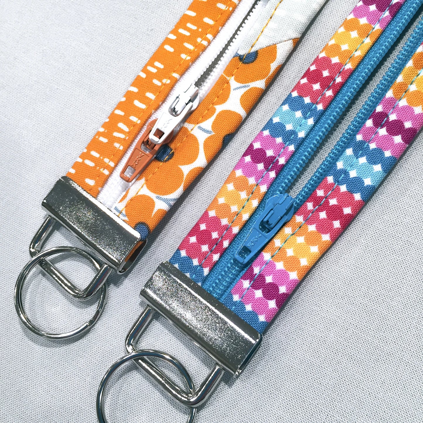 Two fabric zipper keychains laid flat on a light gray textured background. One keychain has an orange and white patterned strap with a silver metal clasp and ring.
[additional_image_link]