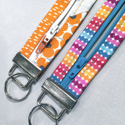 Two fabric zipper keychains laid flat on a light gray textured background. One keychain has an orange and white patterned strap with a silver metal clasp and ring.
[additional_image_link]