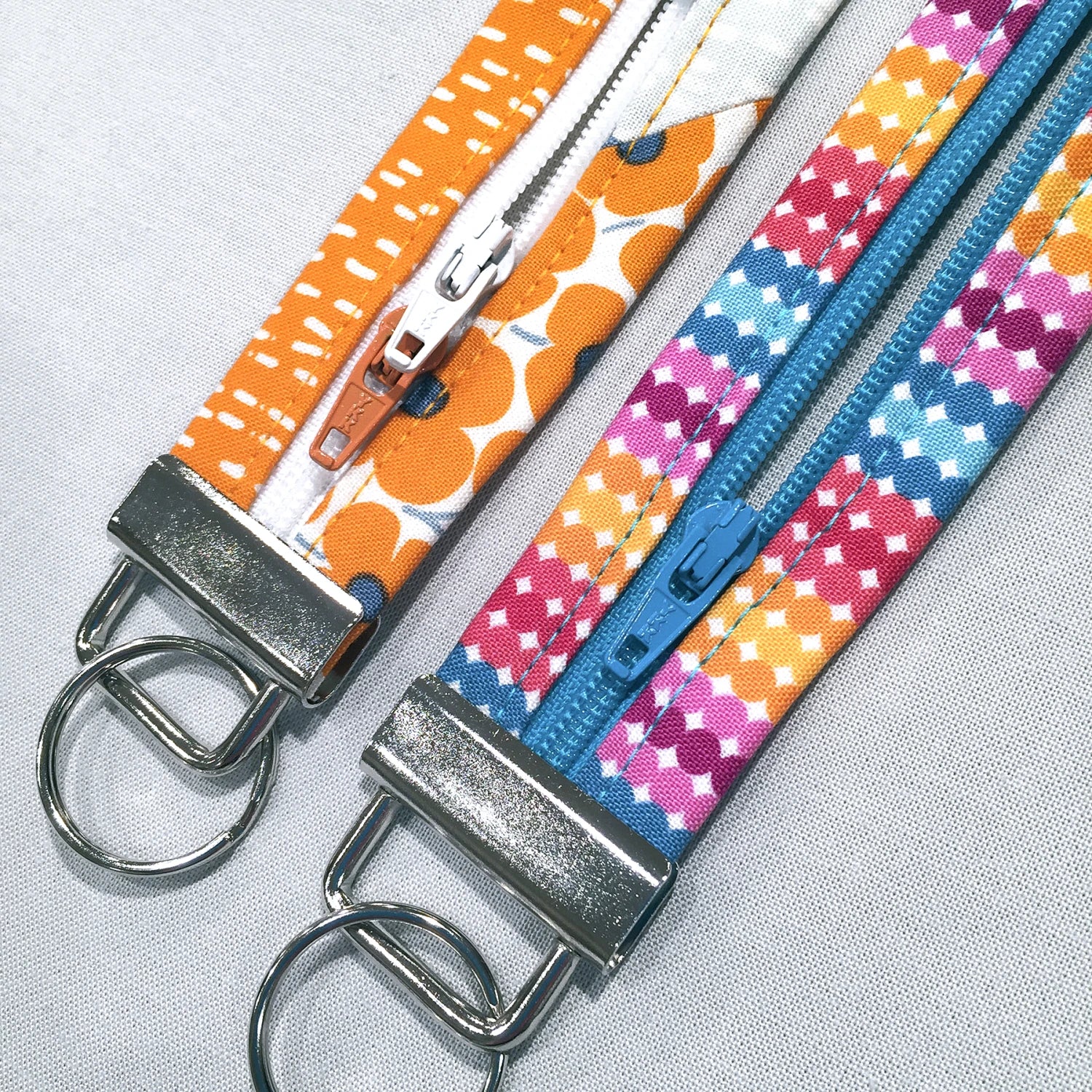 Two fabric zipper keychains laid flat on a light gray textured background. One keychain has an orange and white patterned strap with a silver metal clasp and ring.
[additional_image_link]
