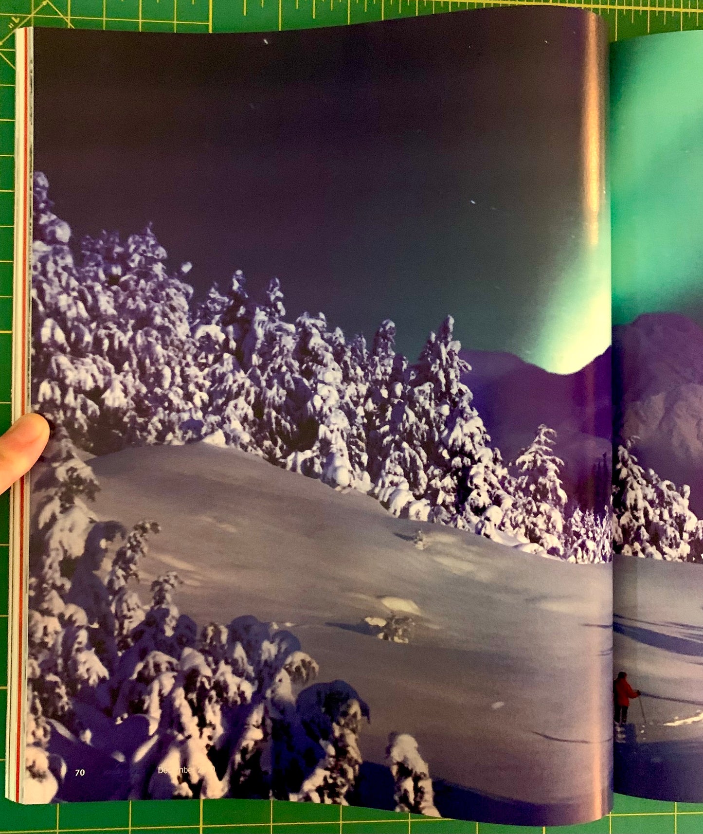 An open book or magazine showing a winter landscape photograph with snow-covered trees, a snowy slope, mountains, and aurora lights. A hand is holding the left edge of the page, and the book is placed on a green cutting mat.
[additional_image_link]