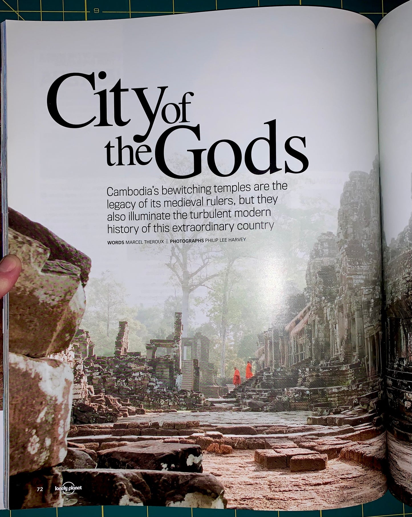 An open magazine or book spread showing a travel article titled "City of the Gods" with a photograph of ancient temple ruins in Cambodia. A hand is holding the left page edge, and the background around the open pages is a green gridded cutting mat.
[additional_image_link]