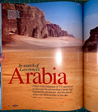 An open magazine or book spread showing a large desert landscape photograph with towering rock cliffs and sandy dunes. The left page has the headline 'In search of Lawrence's Arabia' in large red text, with smaller black and white body text below.
[additional_image_link]