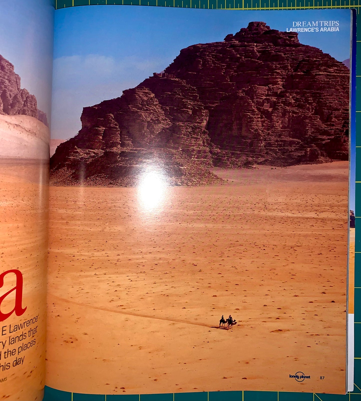 An open magazine or travel book spread showing a desert landscape photograph with a large rock formation, sandy dunes, and a bright sun reflection. The left page has partial text, and the right page includes the headline 'DREAM TRIPS LAWSURGE'S ARABIA' and a small Lonely Planet logo.
[additional_image_link]