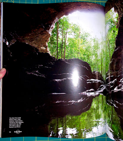 An open book or magazine showing a large scenic photograph of a cave opening over a green forest and reflective pool. A hand is holding the left page edge, and the photo is taken on a green cutting mat.
[additional_image_link]