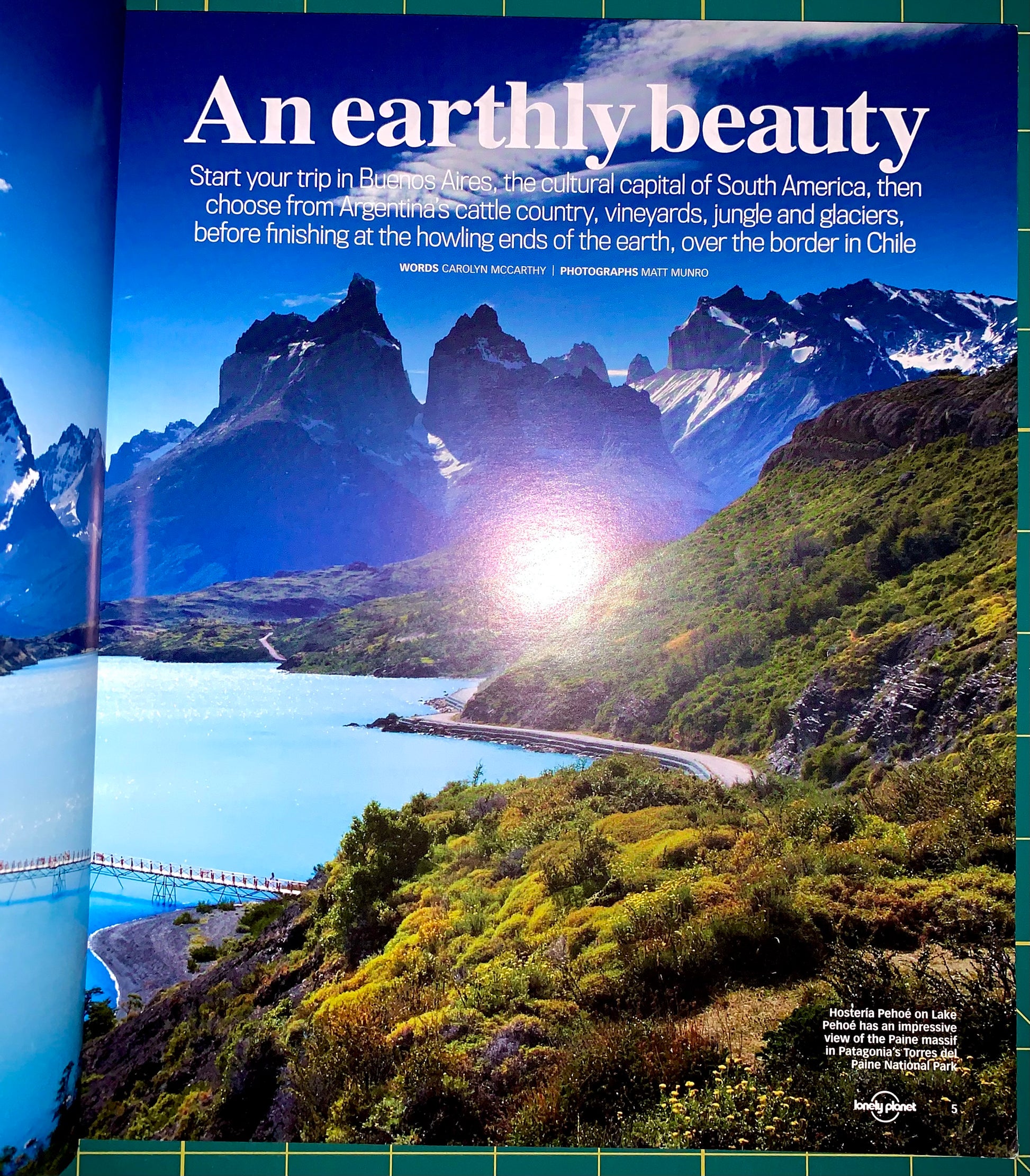 A travel poster or magazine spread featuring a dramatic landscape of a fjord with mountains, blue water, and bright sunlight. Large white headline text reads 'An earthy beauty,' with smaller travel copy above and a small Lonely Planet logo at the bottom right.
[additional_image_link]