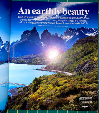 A travel poster or magazine spread featuring a dramatic landscape of a fjord with mountains, blue water, and bright sunlight. Large white headline text reads 'An earthy beauty,' with smaller travel copy above and a small Lonely Planet logo at the bottom right.
[additional_image_link]
