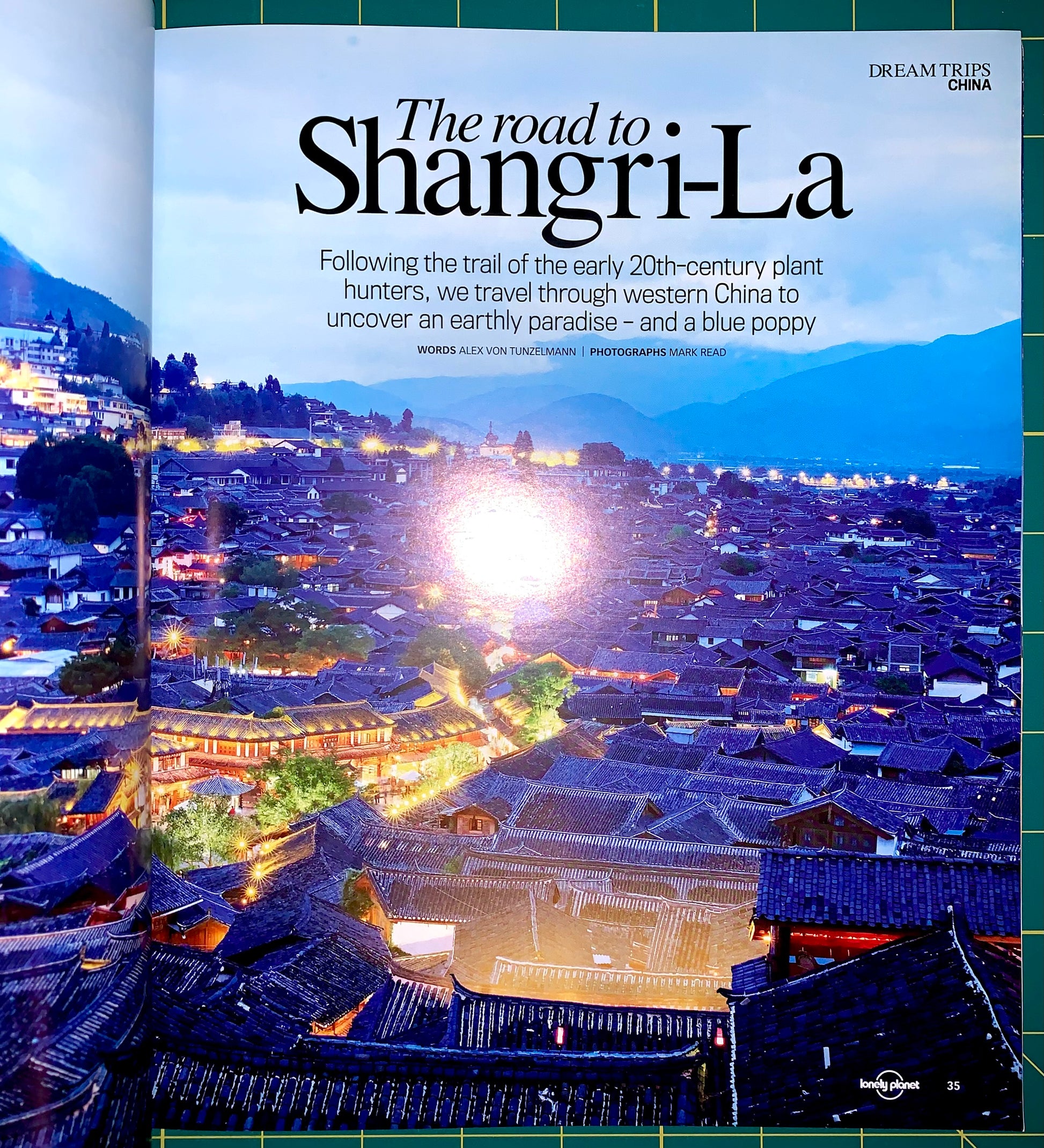 An open travel magazine spread featuring a scenic photograph of a mountain town at dusk with glowing streetlights and large title text reading 'The road to Shangri-La.' The left page shows the photograph and article text, while the right page shows a green grid-patterned cover edge and the Lonely Planet logo near the bottom.
[additional_image_link]