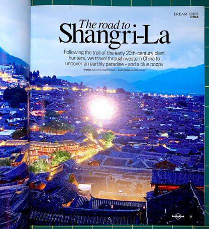 An open travel magazine spread featuring a scenic photograph of a mountain town at dusk with glowing streetlights and large title text reading 'The road to Shangri-La.' The left page shows the photograph and article text, while the right page shows a green grid-patterned cover edge and the Lonely Planet logo near the bottom.
[additional_image_link]