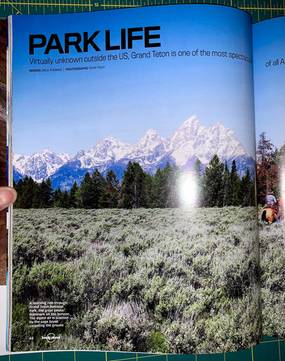 An open magazine or book spread showing a large landscape photograph of snow-capped mountains, evergreen trees, and a sagebrush field under a blue sky, with the headline 'PARK LIFE' at the top.
[additional_image_link]