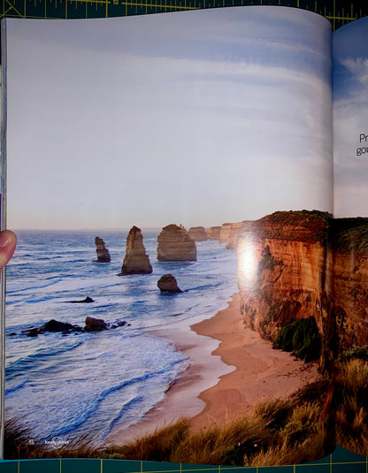 An open book or magazine showing a large coastal photograph of sea stacks, beach, and cliffs, photographed from below on a green cutting mat with a grid.
[additional_image_link]