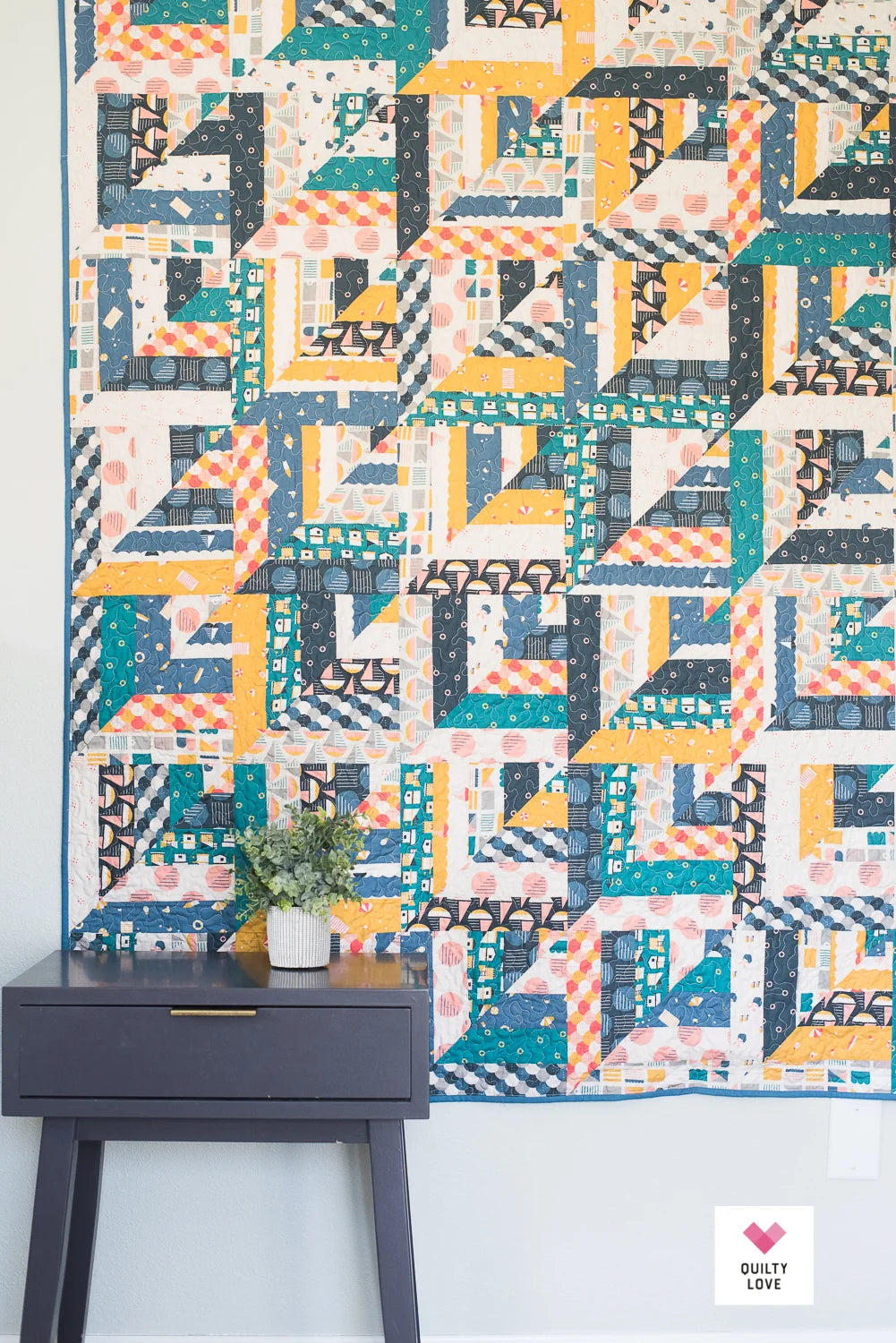A colorful geometric quilt hanging on a wall above a dark gray side table with a small potted plant. The quilt features a patchwork design with repeating blocks of teal, mustard yellow, orange, navy, and cream in various patterned fabrics.
Photo by Quilty Love [additional_image_link]