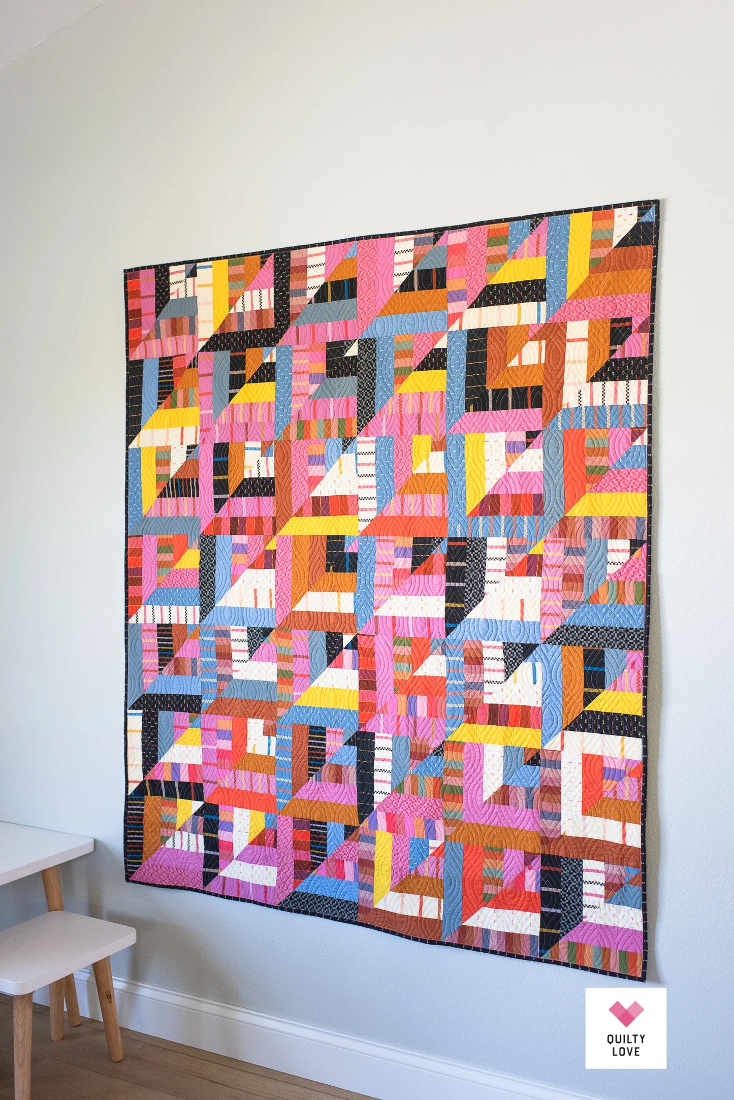 Colorful geometric quilt hanging on a white wall with a small stool in the corner.
Photo by Quilty Love [additional_image_link]