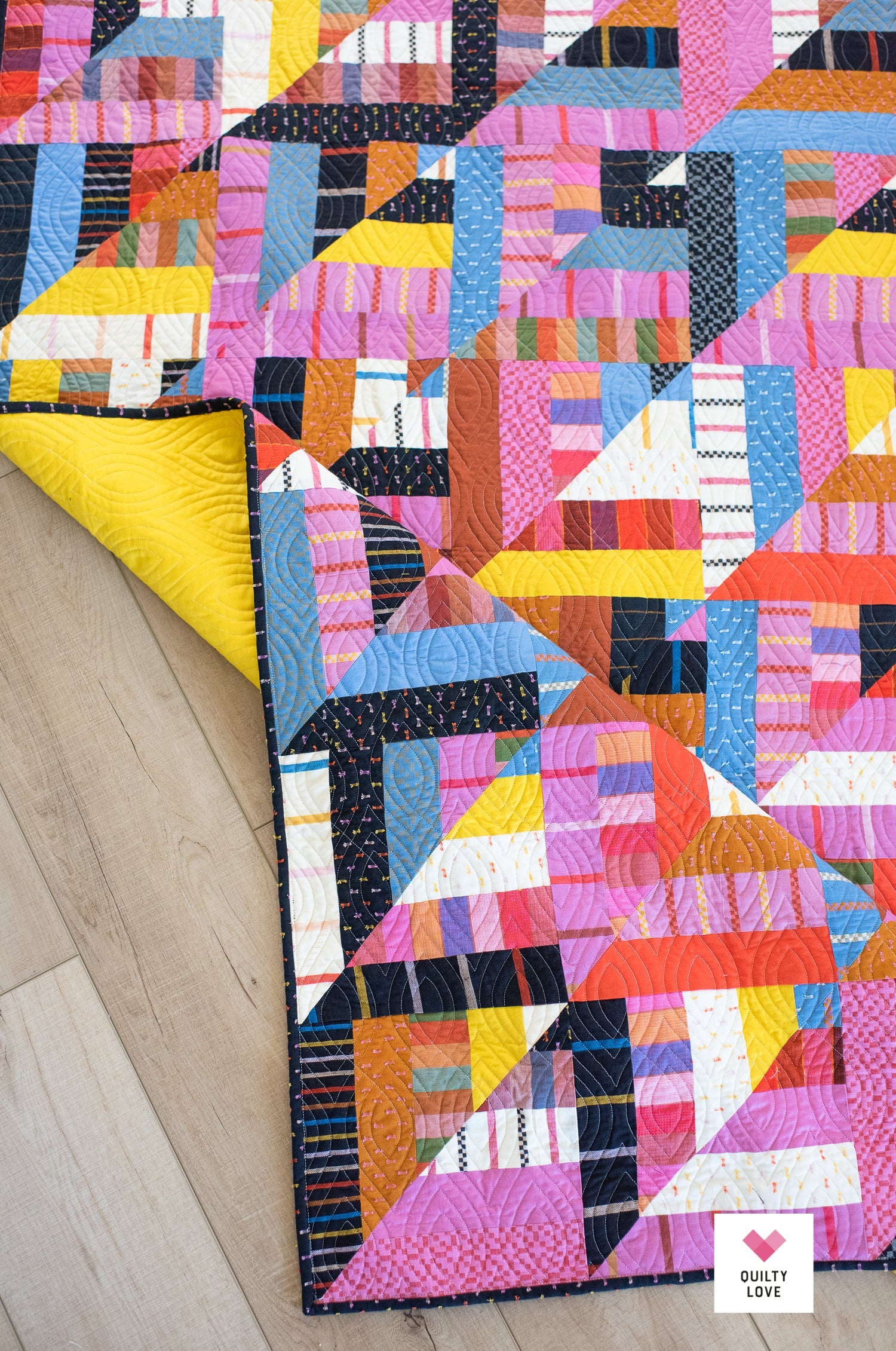 A colorful geometric patchwork quilt laid out on a light wood floor, with one corner folded back to reveal a bright yellow backing. The quilt features a variety of vibrant colors including pink, blue, orange, yellow, and white in a modern triangular pattern.
Photo by Quilty Love [additional_image_link]