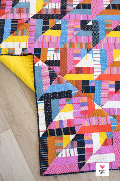 A colorful geometric patchwork quilt laid out on a light wood floor, with one corner folded back to reveal a bright yellow backing. The quilt features a variety of vibrant colors including pink, blue, orange, yellow, and white in a modern triangular pattern.
Photo by Quilty Love [additional_image_link]