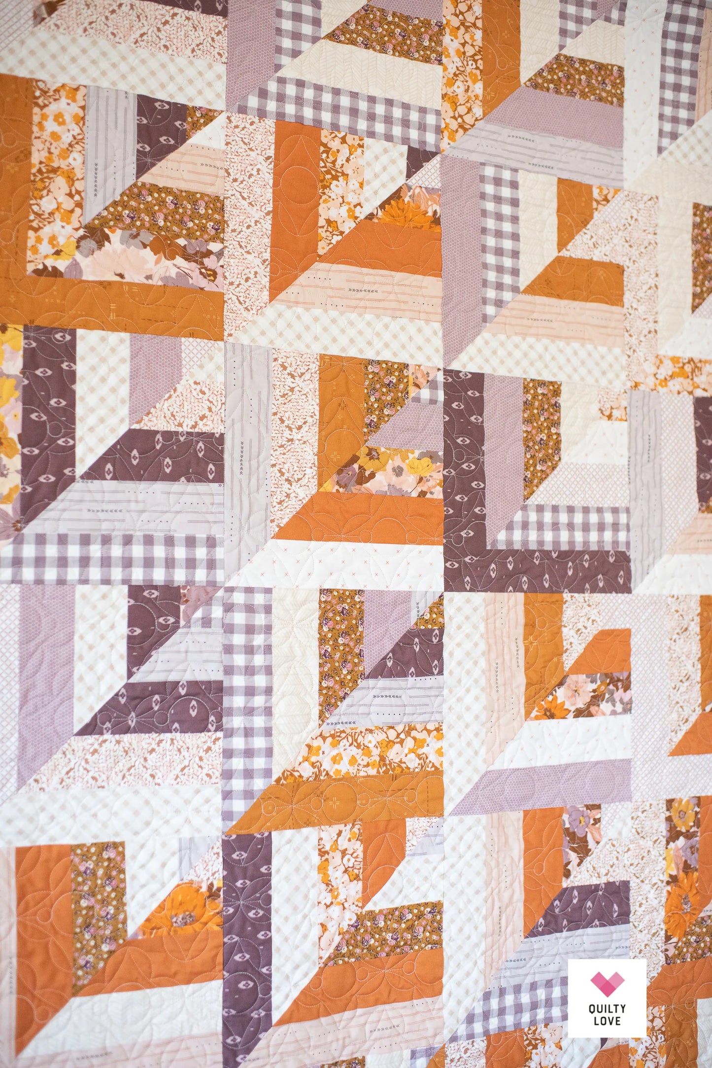 A close-up of a quilted fabric featuring a geometric pinwheel pattern in orange, brown, lavender, and white, with some floral and gingham print fabrics. A small 'Quilty Love' brand logo is visible in the bottom right corner.
Photo by Quilty Love [additional_image_link]