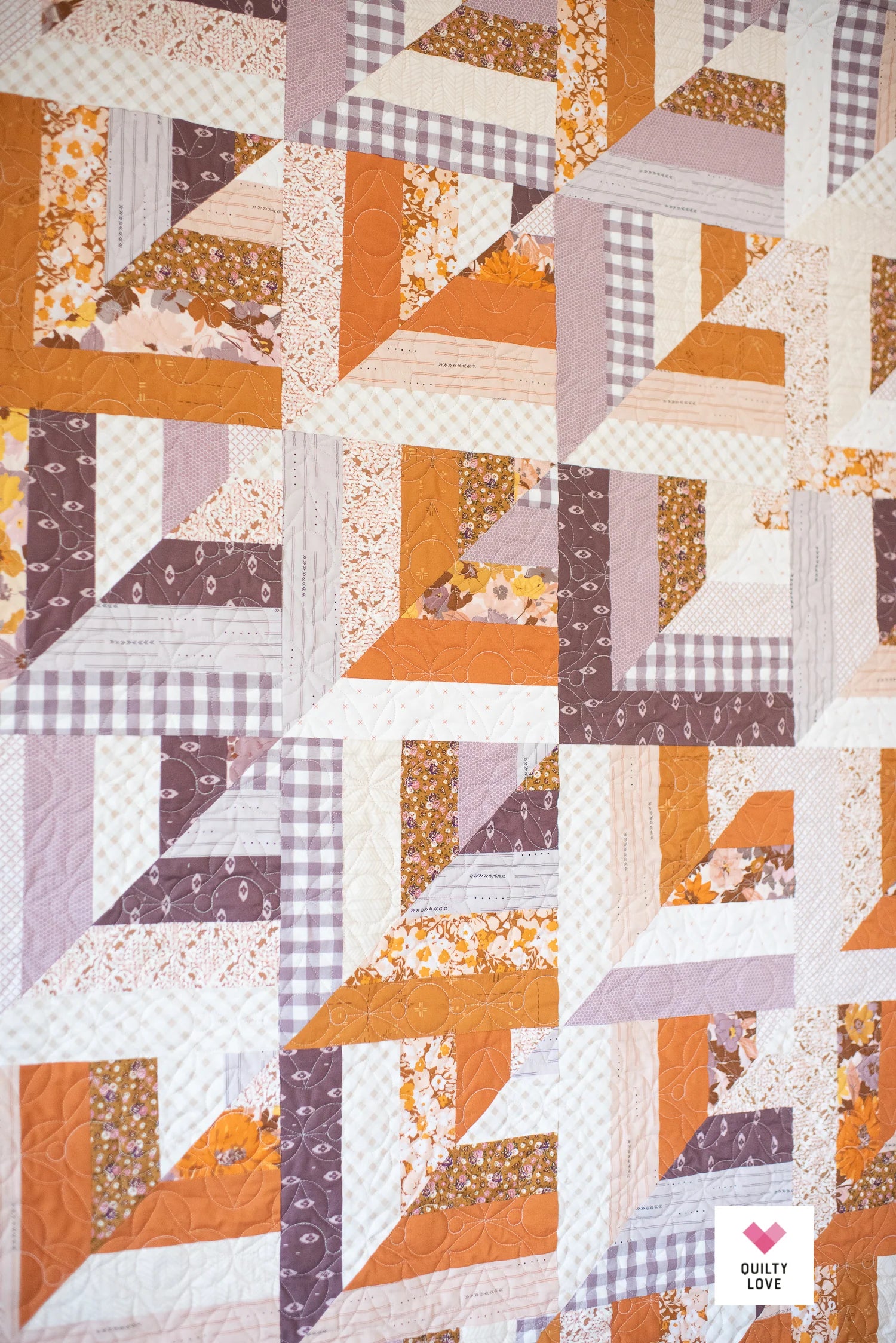 A close-up of a quilted fabric featuring a geometric pinwheel pattern in orange, brown, lavender, and white, with some floral and gingham print fabrics. A small 'Quilty Love' brand logo is visible in the bottom right corner.
Photo by Quilty Love [additional_image_link]