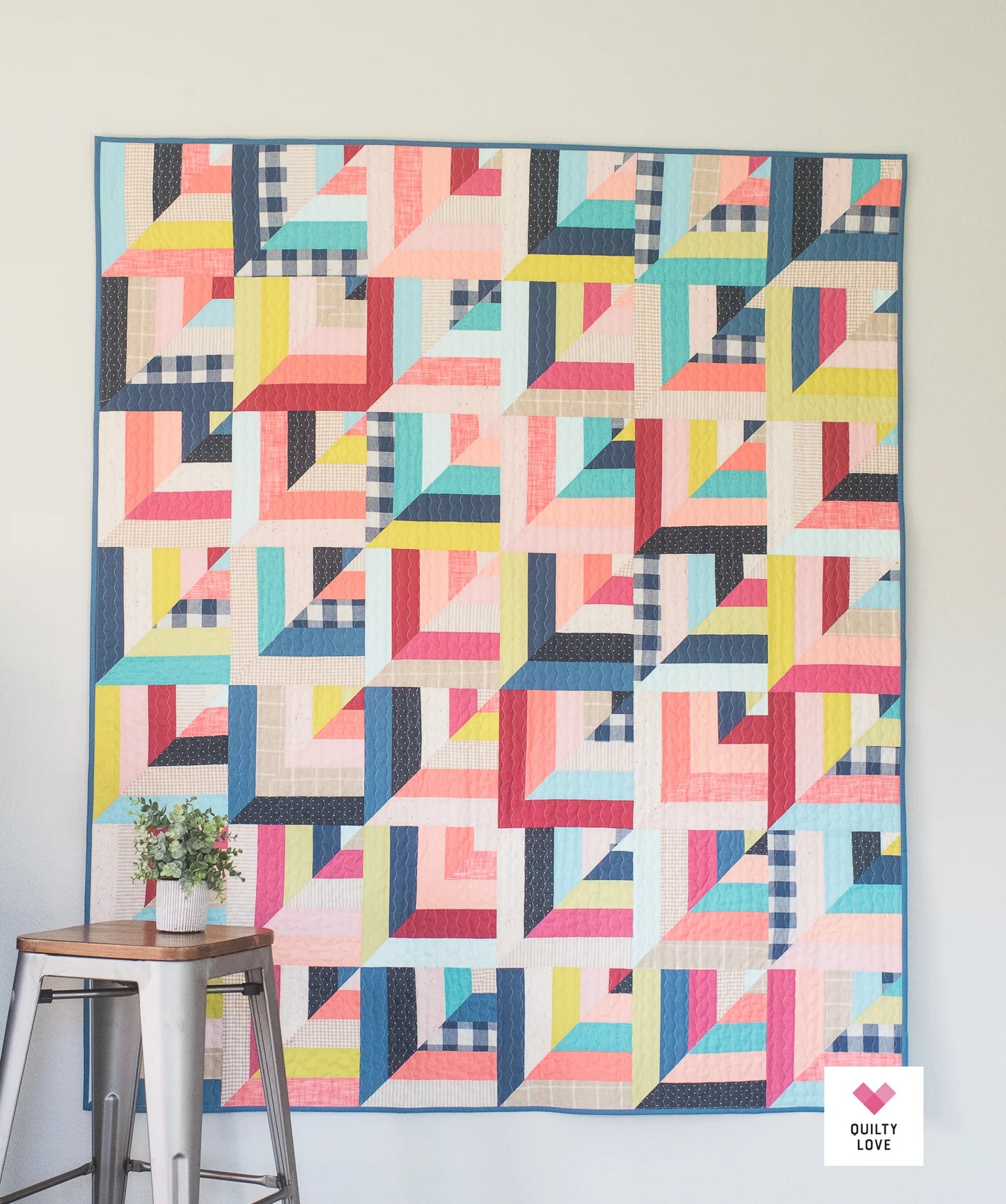 Colorful geometric quilt hanging on a wall with a small plant and stool in the foreground.
Photo by Quilty Love [additional_image_link]