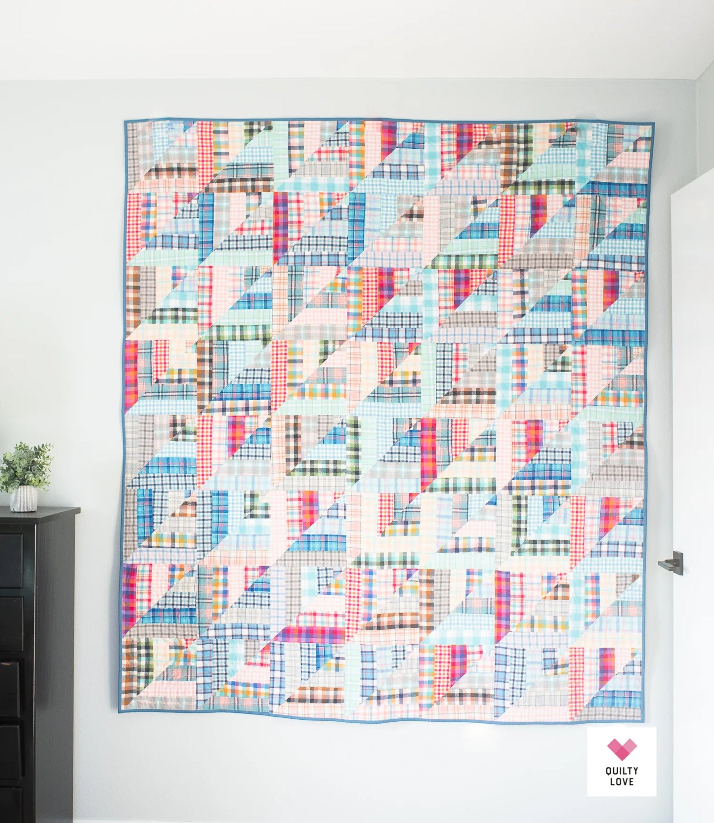 A colorful patchwork quilt hanging on a white wall in a room, featuring a geometric pattern of plaid and striped fabric squares in blue, pink, red, green, and white. A small 'Quilted Love' logo is visible in the bottom right corner.
Photo by Quilty Love [additional_image_link]