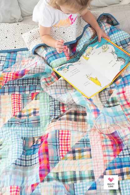 A child sits on a bed reading a colorful children's book, surrounded by a patchwork quilt made of plaid and checkered fabric pieces in pastel and bright tones. A small 'Dusty Love' logo appears in the bottom right corner.
Photo by Quilty Love [additional_image_link]