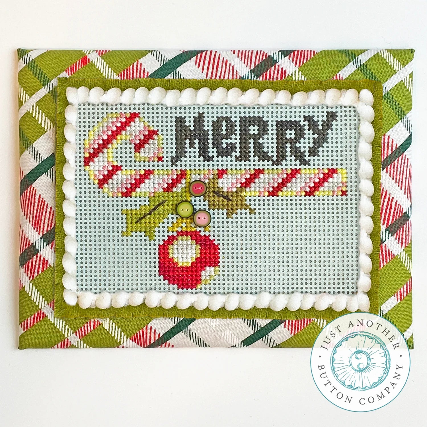 A square handmade cross-stitch holiday card featuring the word 'MERRY' in dark gray, a red ornament, green leaves, and candy canes on a pale blue-gray stitched center. The card is framed with white pearl-like trim and an olive green border, set against a red, green, and white plaid background.
Photo by JustAnotherButtonCompany.com [image_link]