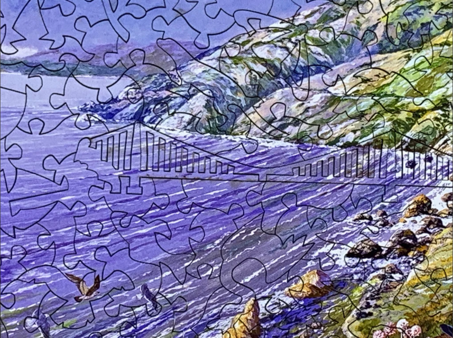 A completed jigsaw puzzle depicting a colorful coastal landscape with purple water, green hills, flying birds, and wildflowers in the foreground.
Photo by Nautilus Puzzles [additional_image_link]