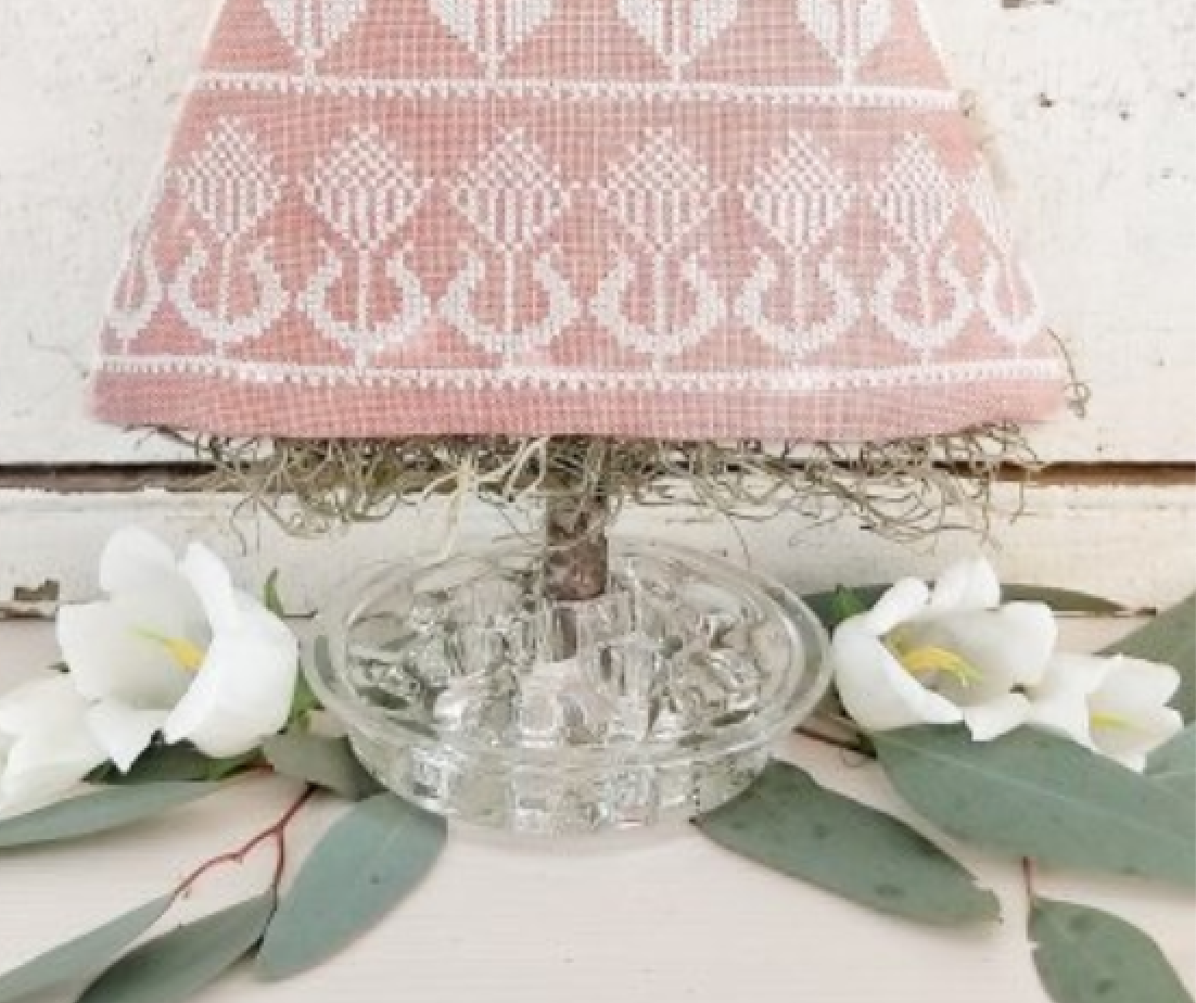 A decorative table lamp with a pink and white embroidered fabric shade, a clear glass base, and a top of green moss, styled with white flowers and eucalyptus leaves on a rustic white surface.
Photo by HelloFromLizMathews.com [additional_image_link]