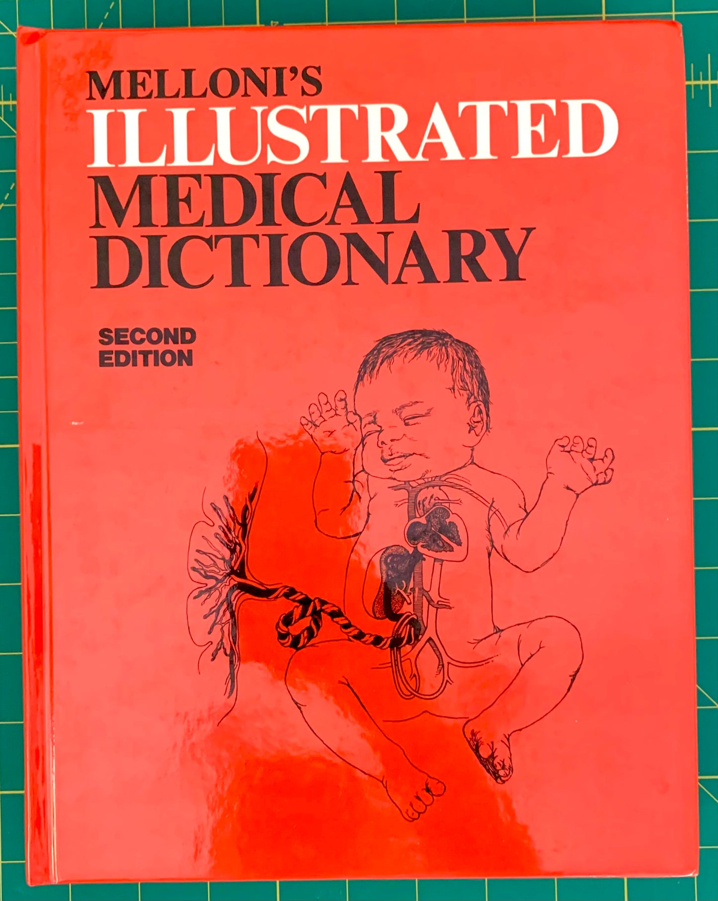 Melloni's Illustrated Medical Dictionary, Second Edition, with a bright coral-red cover, bold black and white title text, and a line drawing of a fetus on the front, photographed on a green cutting mat.
[image_link]