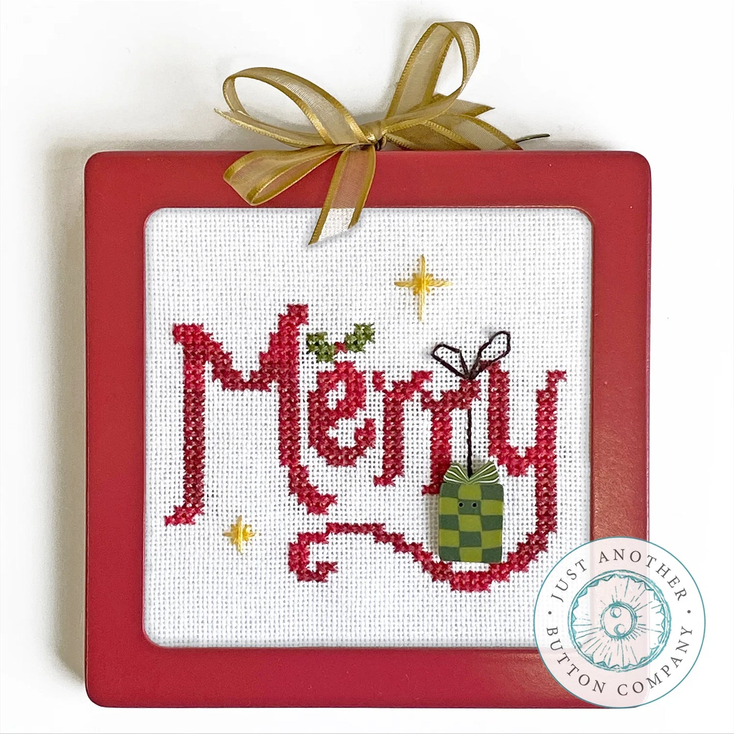 A square red-framed cross-stitch ornament featuring the word 'Merry' in red embroidery on white fabric, with a green gift box, gold stars, and a gold ribbon bow at the top. A circular 'Just Another Button Company' logo is visible in the lower right corner.
Photo by JustAnotherButtonCompany.com [image_link]
