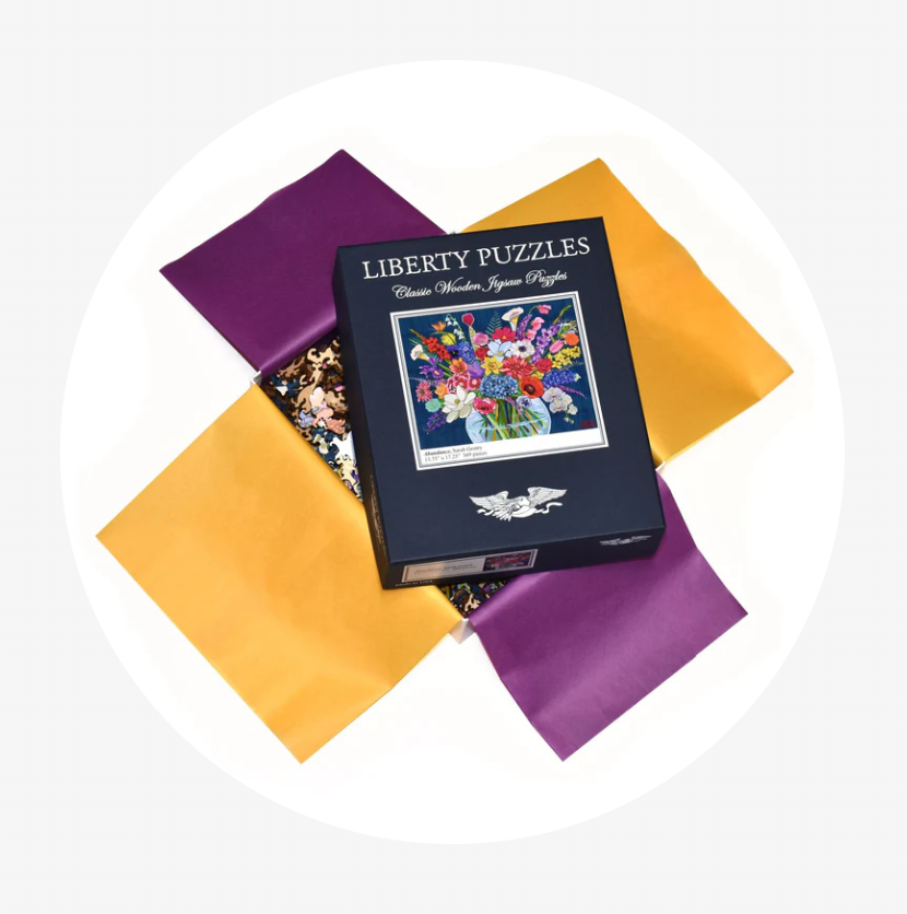 A boxed jigsaw puzzle package labeled 'Liberty Puzzles' sits on top of four satin-like square fabric pieces in purple and mustard yellow on a white circular background.
Photo by Liberty Puzzles [additional_image_link]