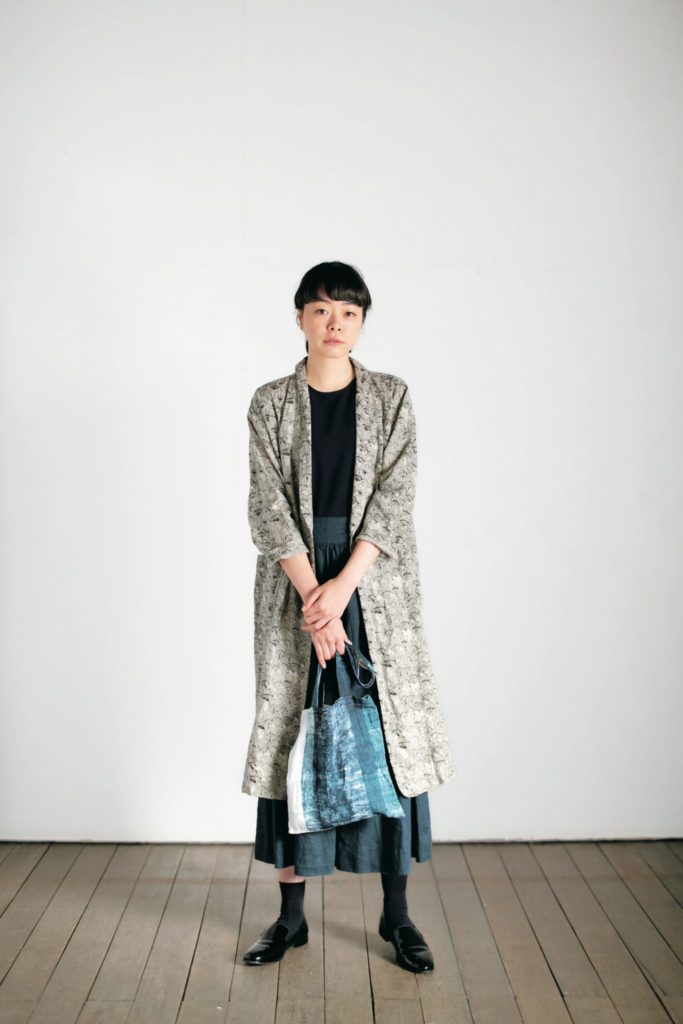 Person wearing a long patterned coat over a dress, holding a bag, on a plain background.
[additional_image_link]