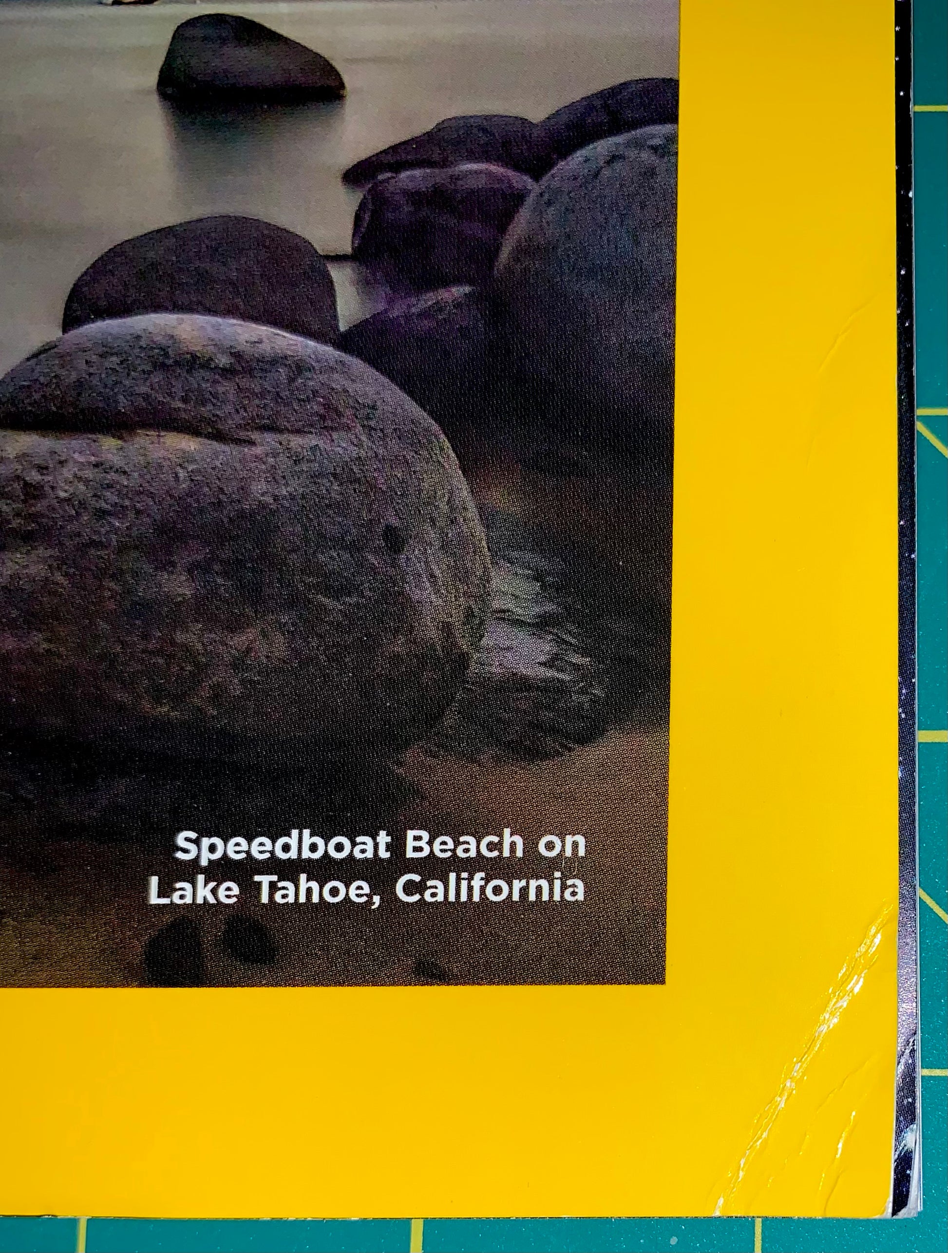 A square print or book cover featuring a photograph of large dark rocks on sandy shore at Speedboat Beach on Lake Tahoe, California, with a bold yellow border and white title text.
[additional_image_link]