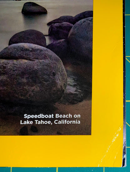 A square print or book cover featuring a photograph of large dark rocks on sandy shore at Speedboat Beach on Lake Tahoe, California, with a bold yellow border and white title text.
[additional_image_link]