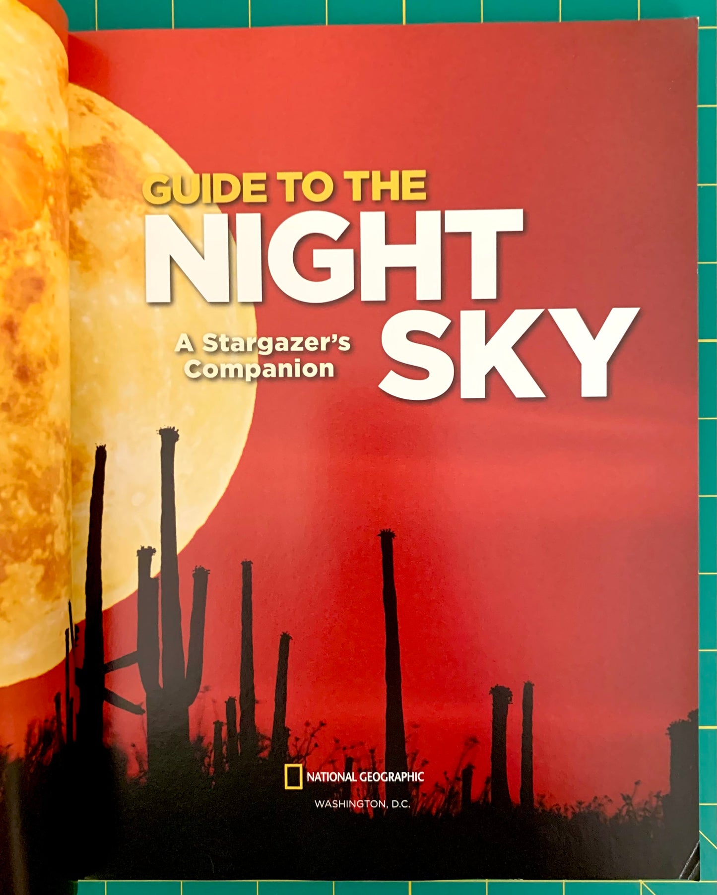 National Geographic Guide to the Night Sky book cover, featuring a large yellow moon against a red sky with silhouettes of saguaro cacti, photographed on a green cutting mat.
[additional_image_link]