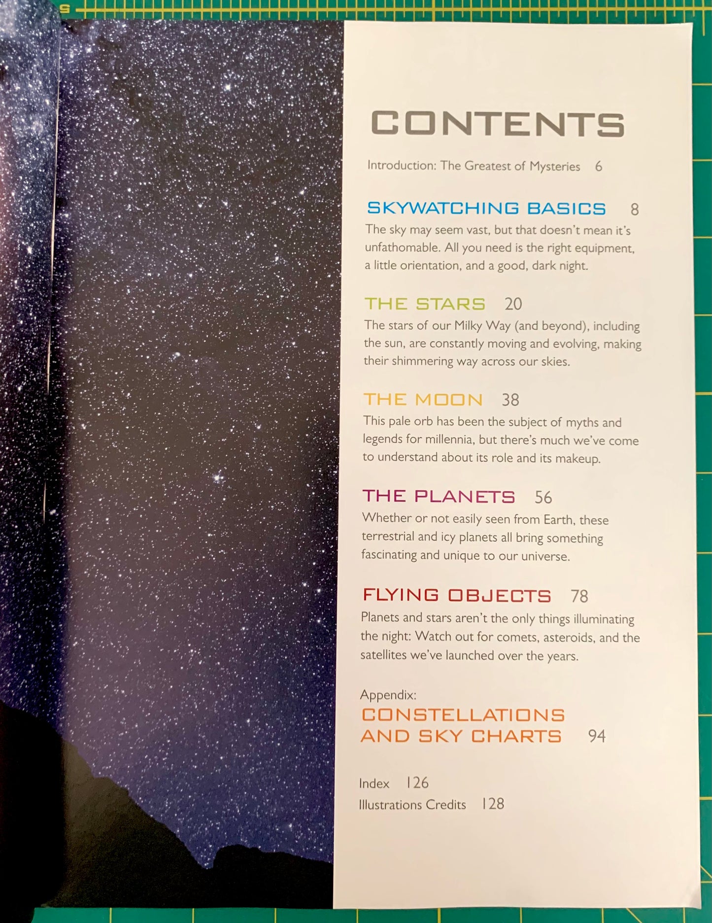 A printed contents page from a book or magazine, showing chapter titles and page numbers in blue and orange text on a cream page, placed on top of a dark blue starry sky photograph. The page is laid on a green grid cutting mat.
[additional_image_link]