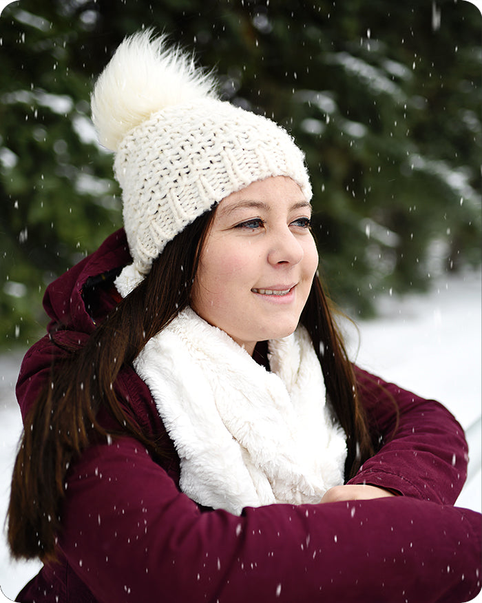 A woman wearing a chunky cream knit beanie with a fluffy pom-pom, a white faux-fur scarf, and a burgundy winter jacket, standing in the snow with trees blurred in the background.
Photo by Shannon Fabrics [additional_image_link]