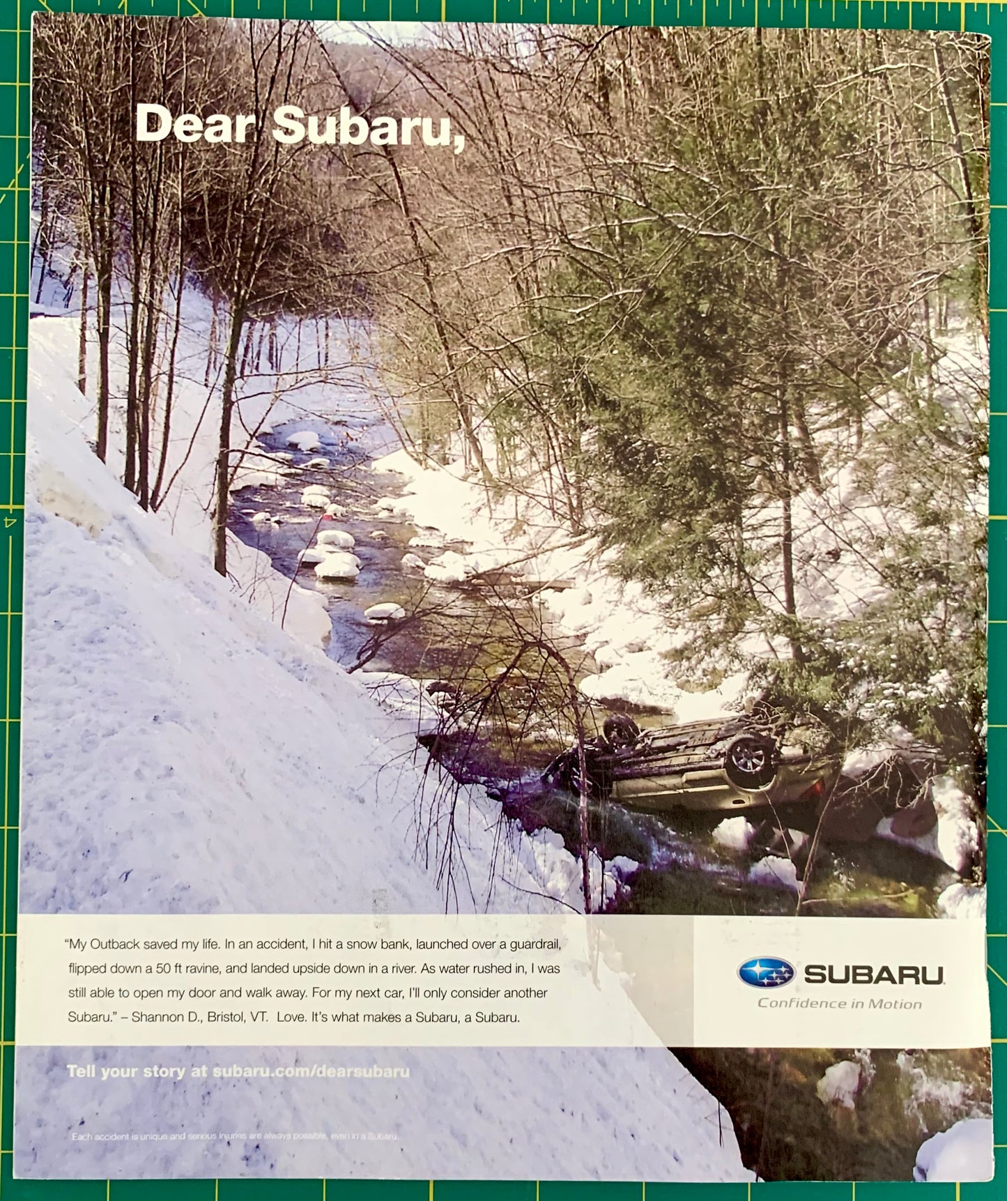 A printed Subaru advertisement or poster featuring a snowy winter forest scene with a stream and an overturned car. Large text reads 'Dear Subaru,' with a Subaru logo and 'Confidence in Motion' at the bottom.
[additional_image_link]