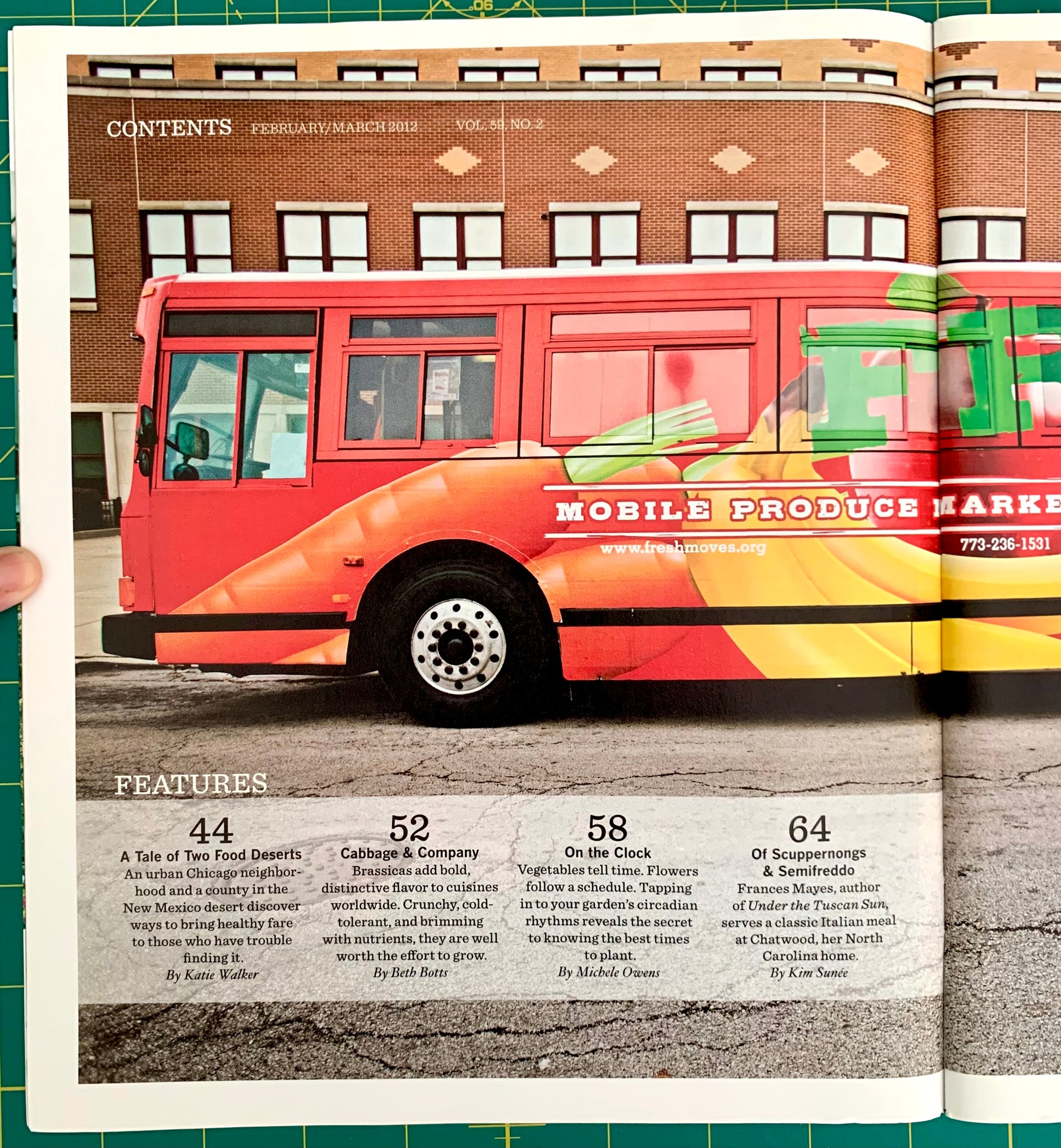 An open printed magazine or catalog spread showing a large red and orange bus advertisement on the left page with text about a mobile produce market, plus a brick building and street scene on the right page. A hand is holding the left page at the left edge.
[additional_image_link]