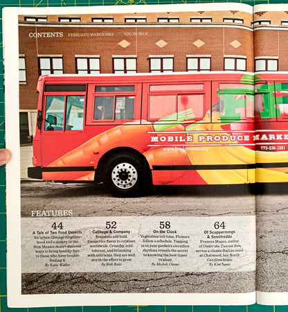 An open printed magazine or catalog spread showing a large red and orange bus advertisement on the left page with text about a mobile produce market, plus a brick building and street scene on the right page. A hand is holding the left page at the left edge.
[additional_image_link]