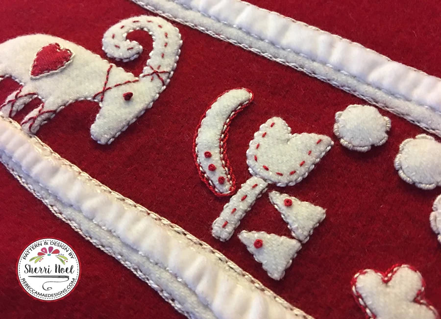 Close-up of a red quilted textile with white appliqué shapes outlined in red stitching, including a stylized animal motif and hearts, bordered by white quilted trim. A circular 'Sherri Noel' watermark appears in the lower left.
Photo by Rebecca Mae Designs [additional_image_link]