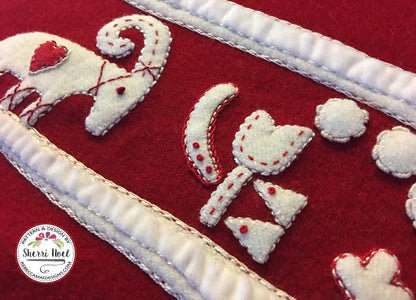 Close-up of a red quilted textile with white appliqué shapes outlined in red stitching, including a stylized animal motif and hearts, bordered by white quilted trim. A circular 'Sherri Noel' watermark appears in the lower left.
Photo by Rebecca Mae Designs [additional_image_link]