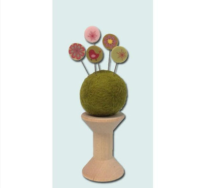 A felt needle holder with a round green wool ball on a light wood stand, holding five colorful flower-shaped pins, shown against a pale blue background. In this kit I substituted a pink felt ball and added the silk ribbon.
[additional_image_link]