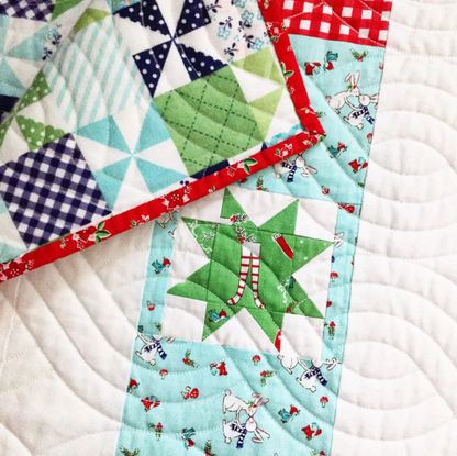 A close-up of a handmade quilted fabric featuring various patchwork designs, including green star sections with candy cane and Santa patterns, red gingham borders, and white quilted background fabric.
Photo by RileyBlakeDesigns.com [additional_image_link]