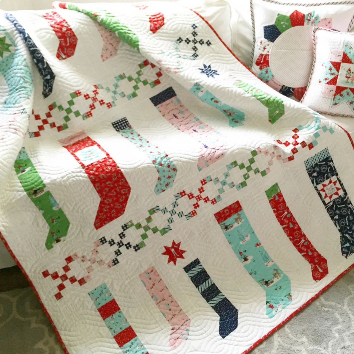 A quilted Christmas-themed blanket or throw draped over a bed, featuring a white background with colorful stitched stocking patterns in red, green, pink, blue, and navy, along with small snowflake and star motifs.
Photo by RileyBlakeDesigns.com [additional_image_link]