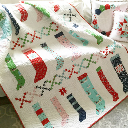 A quilted Christmas-themed blanket or throw draped over a bed, featuring a white background with colorful stitched stocking patterns in red, green, pink, blue, and navy, along with small snowflake and star motifs.
Photo by RileyBlakeDesigns.com [additional_image_link]