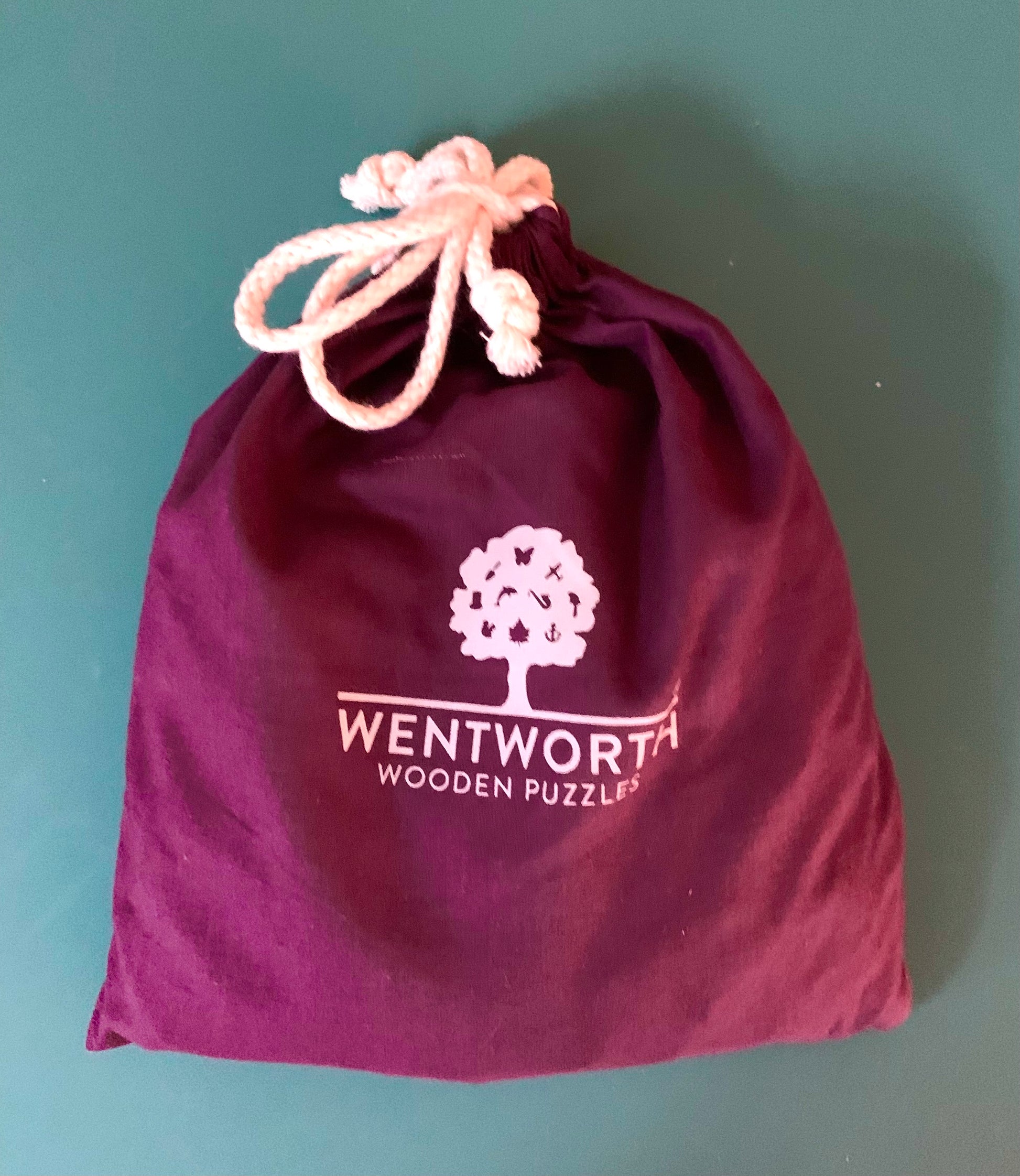 A deep purple drawstring pouch with a white printed Wentworth Wooden Puzzles logo, featuring a light pink drawstring, set against a solid teal background.
Photo by Karen King [additional_image_link]