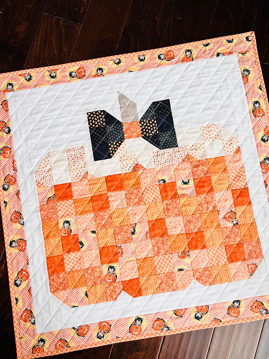 A handmade quilted table runner featuring a pumpkin design in shades of orange, peach, and white, with a patterned border of pumpkins on a dark wood floor.
Photo by My Sew Quilty Life [additional_image_link]