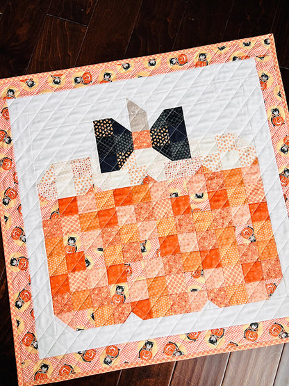 A handmade quilted table runner featuring a pumpkin design in shades of orange, peach, and white, with a patterned border of pumpkins on a dark wood floor.
Photo by My Sew Quilty Life [additional_image_link]