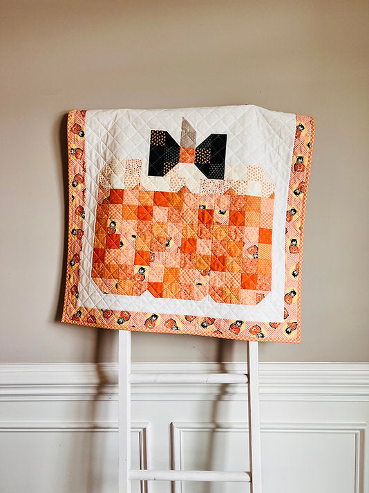 A handmade quilted baby blanket featuring a pumpkin patch design and a black bow motif, draped over a white wooden chair against a beige wall.
Photo by My Sew Quilty Life [additional_image_link]