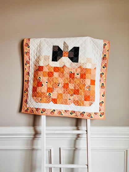 A handmade quilted baby blanket featuring a pumpkin patch design and a black bow motif, draped over a white wooden chair against a beige wall.
Photo by My Sew Quilty Life [additional_image_link]