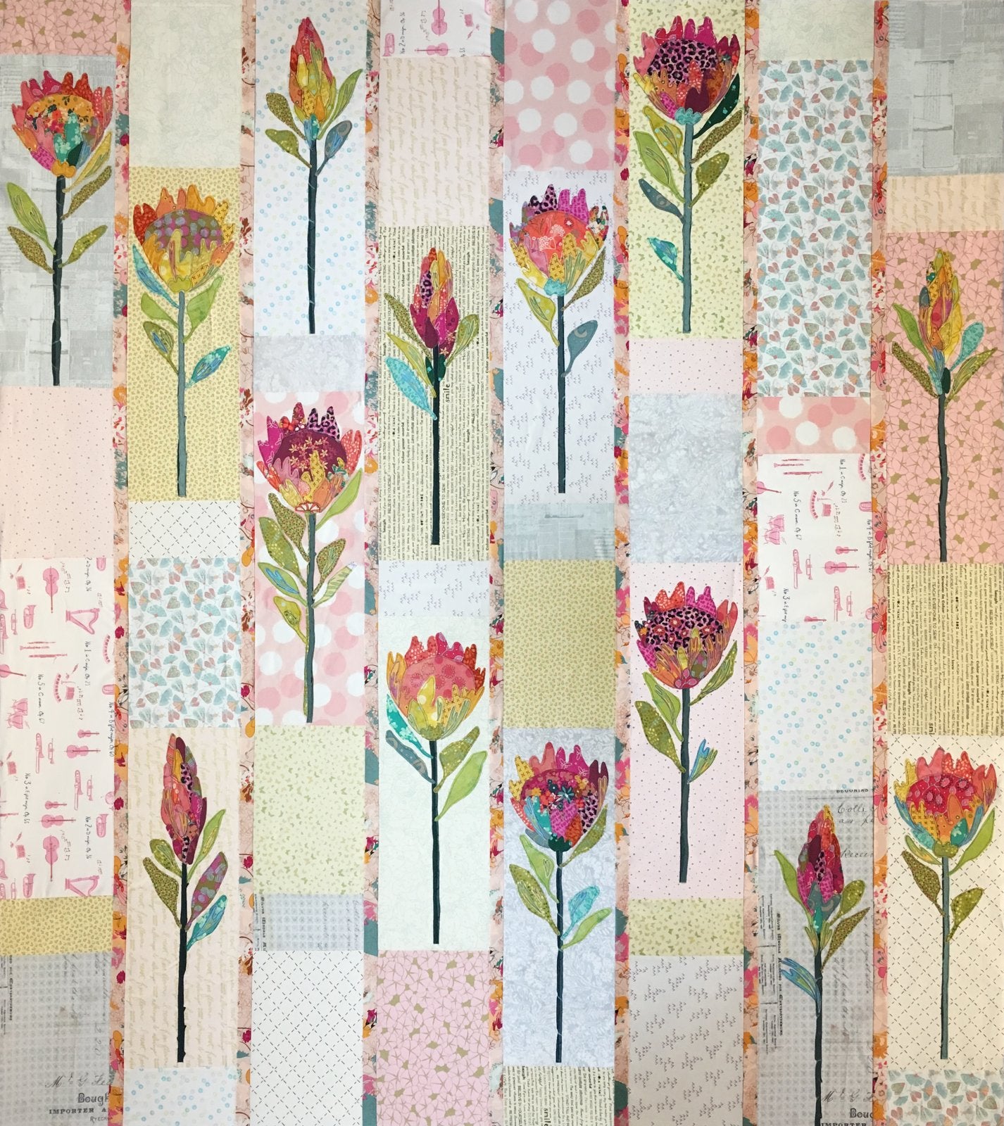 A square patchwork quilt featuring vertical fabric panels in soft pastel colors with embroidered or printed floral motifs, including pink, red, yellow, and green flowers on white, pink, and patterned backgrounds.
Photo by Laura Heine [image_link]